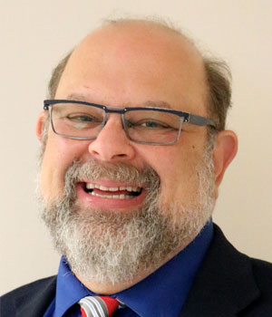 Eric Jacobson, a white man, wears glasses and smiles at the camera. He is bald-headed, with a graying beard and wears a dark suit.